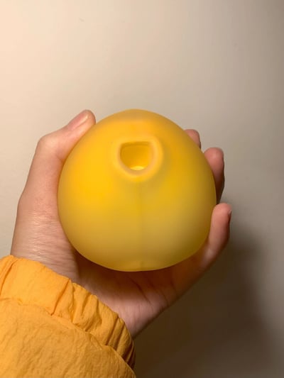 一只手握着一个黄色的磨砂质感圆球，球体呈半透明状，顶部有一个圆形的小孔。手部穿着橙色的长袖衣物，背景是纯色的浅色墙面，光线柔和，突出展示了手中物品的造型和质感。