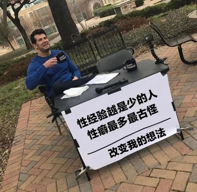 一名男子坐在户外公园的桌子旁，手持黑色杯子，面前的桌子上放着一个写有争议性标语的牌子：'性经验越是少的人 性癖最多最古怪 改变我的想法'。桌子上还放有另一个相同的杯子、麦克风和纸张。男子面前有一个相机三脚架，似乎在进行某种街头采访或社会实验。背景中可以看到公园长椅、树木和建筑物。