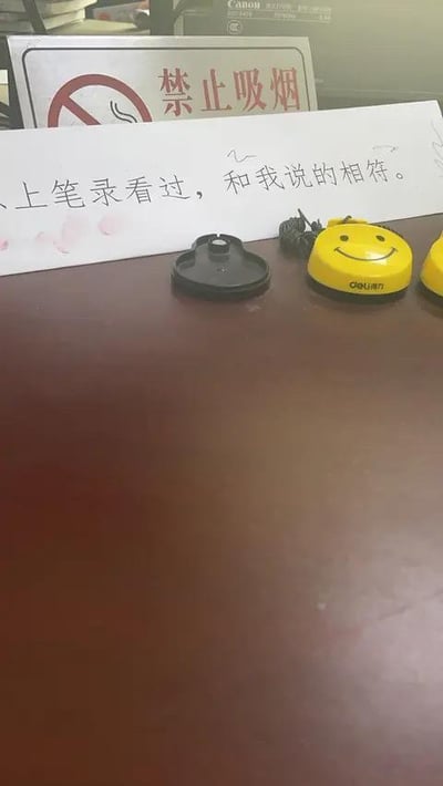 这是一张室内桌面的实拍图，画面里有一张手写纸条，上面写着「以上笔录看过，和我说的相符。」，纸条上还有两处粉色的指印痕迹；纸条后方立着一个标准的「禁止吸烟」标识牌；桌面前景位置有一个黑色的按铃底座，以及两个印有笑脸图案的黄色得力（deli）按铃，背景中还能看到带有Canon标识的设备。场景看起来像是办公或问询类的室内环境，正式的笔录纸条和趣味的笑脸按铃形成了一点反差感。