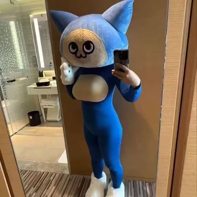 图中是一位穿着原创蓝色卡通猫紧身套装的人在镜子前自拍：套装头部是带有大圆眼睛的呆萌卡通猫脸，手部和脚部为白色造型，身体部分是贴合身形的蓝色紧身衣。此人一只手举着手机对镜拍摄，另一只手做出类似挥手的姿势，背景是带有梳妆台与卫浴区域的酒店风格房间。整体造型将卡通形象与紧身cos服结合，风格滑稽又可爱，充满搞怪趣味。