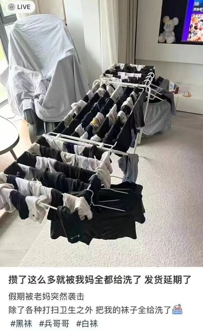 图片展示了一个客厅场景，中央摆放着一个大型折叠晾衣架，上面密密麻麻挂满了各种颜色的袜子（以黑色和白色为主），数量庞大几乎占据了半个房间。背景中可以看到被灰色布单覆盖的椅子、带有米奇玩偶的电视以及浅色地毯。图片下方配有文字，吐槽假期期间被老妈突然袭击打扫卫生，结果攒了很久的袜子全被洗了，导致“发货延期”，充满了年轻人对家长过度关心的无奈又搞笑的吐槽。