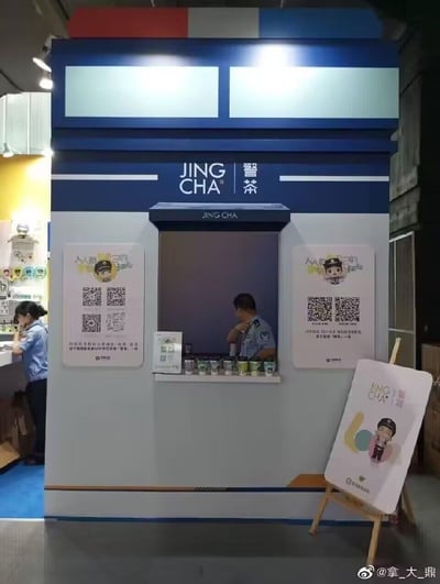 这是一个以谐音梗「警茶」（警察）为核心的创意警务宣传展位，将警务宣传与茶饮店窗口形式结合，展位外观设计成蓝白配色的窗口样式，上方标注「JING CHA 警茶」字样。窗口内有身着警服的工作人员，窗口台面上摆放着茶饮杯；展位两侧张贴带有Q版警察形象的宣传海报，海报上印有「人人都是自己的守护神」标语与二维码，引导观众扫码领取「警茶」。
该设计使用了谐音梗，将「警察」谐音为「警茶」，用贴近大众的茶饮店形式来做警务宣传，形式新颖有趣，降低了宣传的距离感。