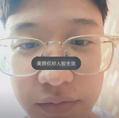 一张近距离拍摄的人物面部照片，人物戴着透明框架眼镜，直视镜头。图片中央有黑色半透明文字框，内写“美颜仅对人脸生效”，暗示美颜滤镜可能无法对眼镜等非面部区域生效的幽默效果。