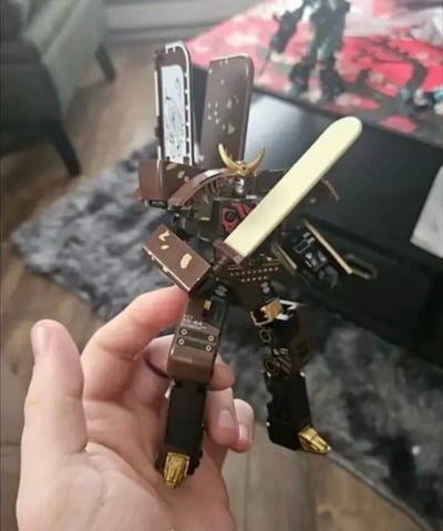 图片中展示了一只手握着一个由电子元件和金属部件手工制作的机器人模型。这个机器人造型独特，具有类似武士的风格，头部有金色的角状装饰，手持一把类似武士刀的长条形部件。机器人主体由各种回收的电子零件构成，包括电路板、金属片和连接器等，呈现出工业风和手工艺术的结合。背景中可以看到房间的一部分，包括桌子和其他物品，暗示这是在家庭环境中拍摄的个人手工作品。