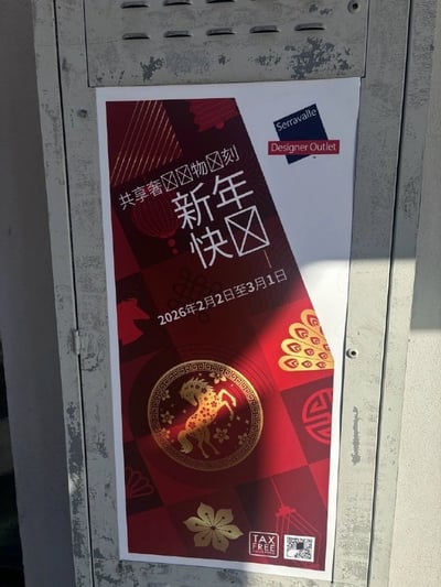 这是一张张贴在灰色磨损金属箱体上的竖版促销海报，是意大利塞拉瓦莱名品奥特莱斯针对2026年中国春节制作的宣传物料。海报整体采用中式新年常见的红金配色，印有金色生肖奔马、传统纹样、桃花、祥云、中国结等春节相关元素，右上角标注品牌标识，底部设有免税标识和活动二维码。海报的中文宣传语存在多处显示异常，汉字被空心方框代替，可识别的内容还包括活动时间为2026年2月2日至3月1日，出现缺字的原因大概率是字体不支持中文导致的显示故障。