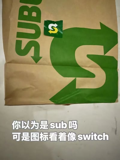 这是一张实拍的赛百味（Subway）牛皮纸包装袋图片，袋子上印有绿色的“SUB”字样与大尺寸循环箭头图案，袋身上方还贴有一个品牌的小尺寸循环箭头贴纸。当贴纸与袋子上的图案组合后，整体视觉效果和任天堂Switch的标志高度相似。图片底部配有白色文字注释：“你以为是sub吗 可是图标看着像switch”，通过这个视觉巧合制造出趣味梗，将快餐品牌元素与知名游戏机标志联系起来，带来幽默的错位观感。