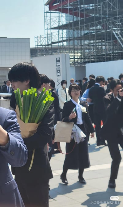 一群身着西装、正装的人聚集在户外，背景可见现代建筑与脚手架，推测可能是毕业礼、公司活动等正式场合。人群中一人格外显眼，他手捧一大束绿色长条状植物（叶片翠绿，顶端有黄色嫩芽），用牛皮纸简单包裹，与周围人的严肃着装形成鲜明反差，画面充满生活气息与趣味性。