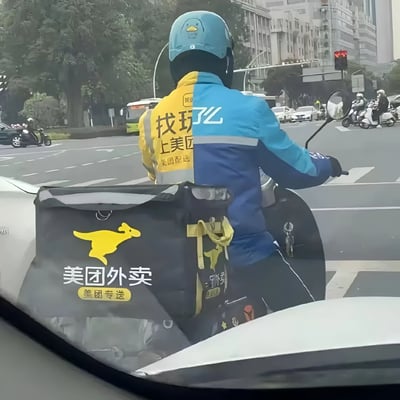图片中是一位正在骑电动车的外卖骑手，他戴着蓝色的卡通鸭头盔，身穿一件左右分色的‘鸳鸯外套’——左侧为美团标志性的黄色，印有‘找玖子上美团 美团专送’字样；右侧为蓝色。车后座上是印有黄色袋鼠logo和‘美团外卖 美团专送’字样的黑色外卖箱。背景是城市街道，有其他车辆和行人，整体场景展现了外卖骑手的日常工作状态，而独特的分色外套设计显得格外吸睛有趣。