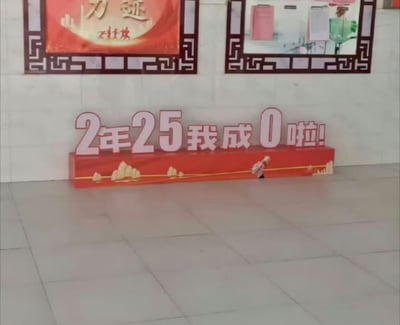 这是一张室内场景的实拍图，地面上摆放着一个带有红色底座的立体标语牌，牌面醒目地写着“2年25我成0啦!”。标语牌底座右侧有一个小型玩偶，通过绳子与底座相连，视觉上仿佛是这个玩偶将数字“25”转变为“0”，让原本偏向正式的成果表述变得充满趣味。背景墙面上设有两个带中式传统边框的装饰牌，其中左侧红色装饰牌上印有“力迹”和“石库大楼”的字样。
该标语大概率是用于宣传某项成果，比如在两年时间内将原本为25的安全事故数、失误次数等指标降至0，借助创意的视觉设计让这一成果更具传播性。