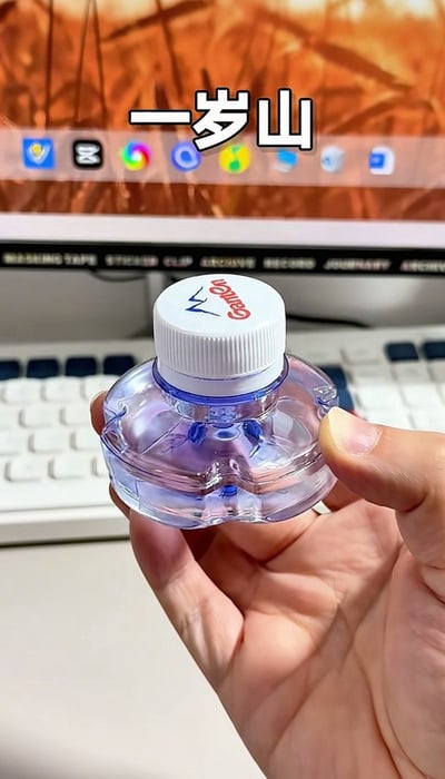 图中一只手正举着一瓶造型和普通百岁山矿泉水一致但尺寸极小的迷你款景田矿泉水，背景为办公场景的电脑显示器与键盘，图片顶部配文“一岁山”，利用谐音和尺寸反差玩梗：原本的矿泉水品牌名为“百岁山”，这款迷你款的尺寸被网友调侃对应“一岁”，因此得名“一岁山”，趣味感十足。