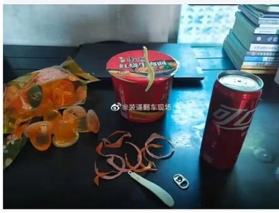 这张图呈现了一个搞笑的生活失误场景：深色桌面上摆放着康师傅红烧牛肉桶面、可口可乐罐和一袋拆开的果冻，桶面的叉子还插在桶盖上，旁边却有被撕成环状的叉子塑料包装，可乐罐的拉环则被取下放在一旁。显然是当事人想要打开可乐时，误把泡面叉子的塑料包装当成了可乐拉环来撕，闹出了令人哭笑不得的小笑话，充满了生活的尴尬趣味。