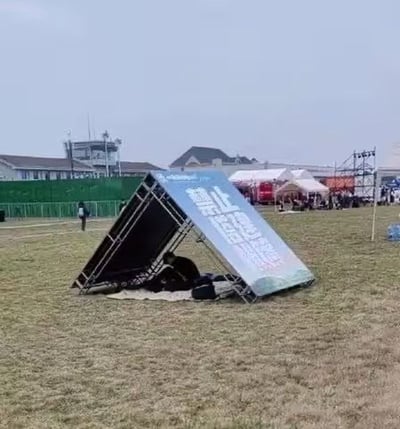 这是一张户外实拍照片，拍摄于某户外活动的草地场地，地面是偏枯黄的草皮。一块带金属桁架的大型宣传广告牌歪倒在地面，倾斜的牌面与地面形成了三角形的遮蔽空间，有人员躲在这个空间里休憩，将倒伏的广告牌当作临时遮阳棚使用。广告牌上印刷的文字因倒伏呈现反向，正向内容为“停止内卷 立刻躺平”，与下方人员躲在阴凉处休息的状态恰好形成呼应，趣味十足。远处可见活动现场的其他帐篷、聚集的人群、绿色围栏，更远处是几栋民用建筑，天空呈灰蒙蒙的阴天状态。