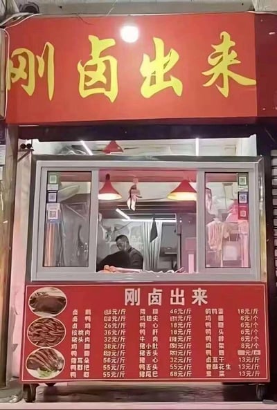 这是一张合成的搞笑梗图，展示了一家名为“刚卤出来”的卤味店：店铺上方是红底黄字的“刚卤出来”招牌，下方是红底白字的价目牌，罗列了卤牛肉、卤鸡胗、鸭头等数十种卤味的价格。但透过卤味店的窗口，内部场景却不是制作卤味的后厨，而是一位理发师正在给顾客理发的理发店场景，形成了强烈的错位反差。店名“刚卤出来”也因此产生了双关趣味，仿佛在调侃顾客“刚被‘理（卤）’出来”，整体画面荒诞又搞笑。