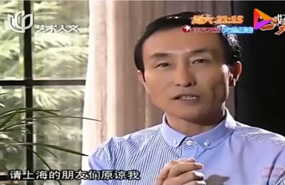 这是一张电视采访截图，画面中一名穿着蓝色条纹衬衫的男子双手合十，表情诚恳。屏幕底部有字幕：'请上海的朋友们原谅我'。该图与'沪国'梗相关，'沪国'是网民对上海的戏谑性称呼，暗示其特殊性如同独立国家。