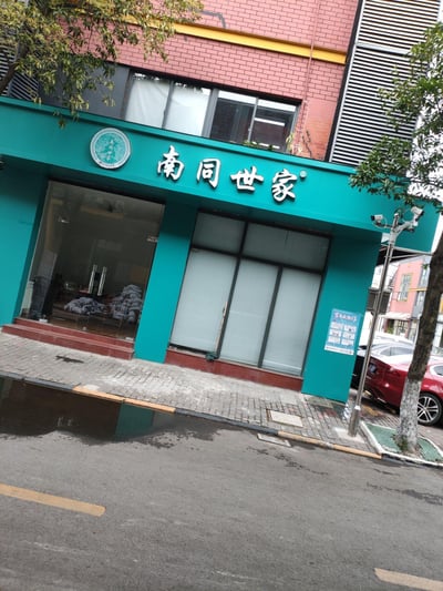 这是一张街景实拍照片，展示了「南同世家」的线下门店。店铺门面采用青绿色设计，上方配有白色的品牌名称“南同世家”，左侧还有圆形品牌标识，标识内标注有“南同世家 始于1618年”字样。玻璃门内可见堆放的白色袋状物，旁侧窗户装有百叶窗。店外人行道有积水痕迹，旁边生长着树木，还有监控杆与一辆停放的红色轿车，背景是红砖结构的多层建筑，整体呈现出日常的城市街景风貌。