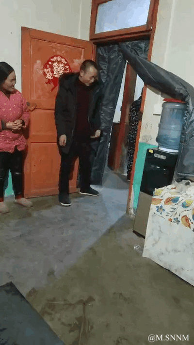 这是一段记录生活日常的动态图，画面呈现的是一个室内门口场景，木门上贴着红色的喜字，一位身着黑色外套的男子正迈步走进屋内，门口的深色门帘因垂落角度恰好与男子的头部位置重合，形成了视觉错位效果，看上去仿佛门帘套在了男子的头上，旁边穿着粉色带点上衣的女子正笑着看向男子，整个场景充满了生活化的意外趣味，是偶然出现的搞笑视觉错觉瞬间。