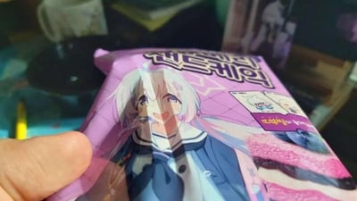 这是一张日常实拍的静态图，画面中一只手拿着一款紫色的零食包装，包装上印有二次元手游《蔚蓝档案》中的角色空崎白子——她有着浅白色长发、蓝紫色眼眸，面带灿烂笑容，身着蓝白水手服。包装上带有韩文标识，翻译后可知这是一款夹心蛋糕类零食，右下角展示了零食的样貌，是带有粉色夹心的点心。背景处于虚化状态，能隐约看到桌面的一些物品，整体氛围轻松可爱，是二次元游戏与零食品牌的联动周边实拍。