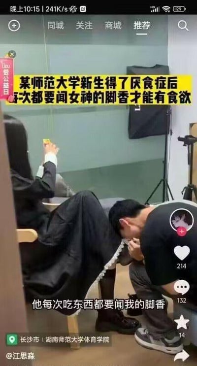 图片显示一名男性学生跪在地上，低头靠近一名坐在椅子上的女性的脚部，似乎在嗅闻。图片上方有黄色大字标题称“某师范大学新生得7厌食症后每次都要闻女神的脚香才能有食欲”，下方配有文字“他每次吃东西都要闻我的脚香”。场景疑似在室内房间，背景有玻璃墙和摄影设备。该内容明显属于低俗猎奇类虚假信息，利用耸人听闻的标题吸引眼球。
