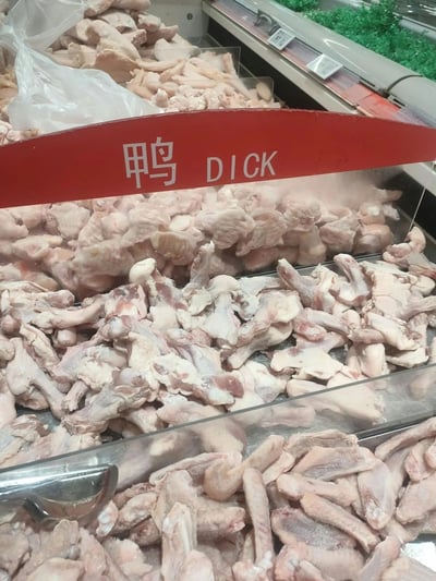 超市冷柜区的生鸭肉陈列场景，红色标牌上用中文写着'鸭'，下方却错误地标注了英文'DICK'（英文中为粗俗词汇，正确翻译应为'duck'）。冷柜中堆满了切块的生鸭肉，背景可见其他肉类和绿色蔬菜陈列。这一明显的翻译错误产生了幽默效果，属于典型的生活场景中的搞笑乌龙。