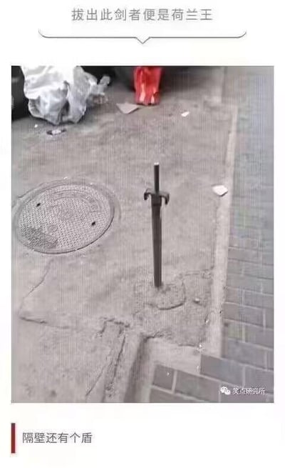 这是一张街头实拍的趣味照片，地面的水泥里固定着一根形似剑柄的金属杆，看起来就像一把插在地里拔不出的剑。图上方配文“拔出此剑者便是荷兰王”，下方配文“隔壁还有个盾”，将旁边的圆形井盖调侃成是配套的盾，模仿西方传说中“拔出石中剑成为王者”的经典设定，把普通的街头设施进行趣味化解读，制造出荒诞搞笑的效果。
