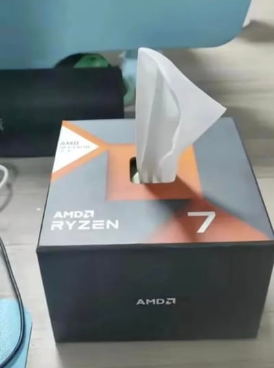 图片展示了一个被创意改造成纸巾盒的AMD Ryzen 7 CPU包装盒。黑色的包装盒上印有白色的“AMD RYZEN”字样和数字“7”，顶部被切割出一个开口，一张白色纸巾正从开口中抽出。背景中可见模糊的黑色物体（疑似音箱）和蓝色物品，整体场景像是在桌面环境中。这是科技爱好者将废弃CPU包装盒二次利用的DIY作品，兼具实用性与创意趣味。