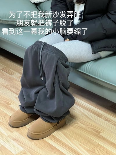 图片中一人坐在浅蓝色沙发上，穿着黑色羽绒服、灰色紧身裤和棕色雪地靴，灰色运动裤被拉至脚踝处，露出里面的紧身裤。图片上方配有文字：“为了不把我新沙发弄脏 朋友就把裤子脱了 看到这一幕我的小脑萎缩了”，展现了朋友为保护新沙发而做出的夸张举动，带有搞笑和无奈的氛围。
