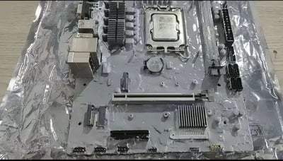 这张图片展示了一块经过手工改造的电脑主板，主板被包裹在透明防静电塑封袋中，放置在浅色木纹桌面上。整个主板的PCB板材和所有电子元件都被统一涂上了银色颜料，原本的PCB颜色、电路印刷标识都被完全覆盖，仅CPU插槽的保护盖上还能隐约看到原有的刻字，图中可以清晰分辨出CPU插槽、PCIe显卡插槽、供电接口、CMOS纽扣电池、散热片等常规主板结构，是一件非常另类的硬件改造作品。