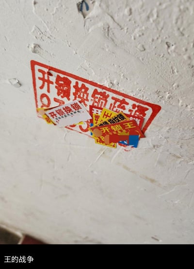 这张图拍摄了墙面上的开锁广告场景：一个大的红底“开锁换锁疏通”印子上，被多张更小的开锁类广告覆盖，这些小广告包含“开锁换锁 保险柜配汽车钥匙”“开锁王”“指纹锁”等不同商家的宣传内容，图片底部配文“王的战争”，用幽默的方式调侃开锁商家之间互相覆盖广告的竞争行为，将现实里的小广告乱象趣味化为一场“领地争夺”的戏码。