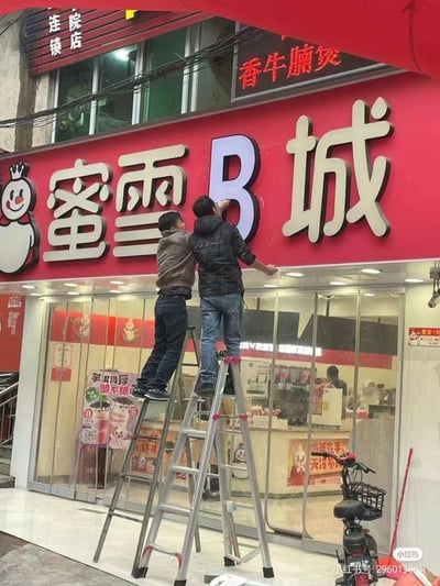 这张图实拍了两个工人站在梯子上，将蜜雪冰城门店招牌上的“冰”字替换成了字母“B”，原本的“蜜雪冰城”变成了“蜜雪B城”，玩了谐音梗，画面充满滑稽搞笑的氛围，让人忍俊不禁。
谐音梗说明：将“冰（bīng）”替换为发音相近的字母“B”，制造出恶搞的趣味效果，调侃知名奶茶品牌蜜雪冰城。