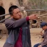 图片中前景是一名身穿灰色外套、内搭红色衣物的男子，他身体前倾，双手紧握一根长木棍，表情用力，似乎在进行某种劳作。背景中有一位戴蓝色帽子的人坐着，周围是土黄色的窑洞式建筑和一些农具，整体场景呈现出典型的乡村或山区生活氛围。