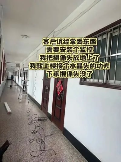 这张图拍摄于一处室内长走廊，是监控安装施工现场，地面散落着黑色的监控网线，墙边立着人字梯，近处的门上贴着福字，整体是普通单位或居民楼的走廊场景。图上叠加了黄色描黑边的文字，讲述了一件充满戏剧性反差的搞笑乌龙事件：客户因为经常丢东西，联系工人上门安装监控，工人把新摄像头临时放在地上，仅仅上楼接水晶头的一小会儿，下楼就发现摄像头已经被偷走了，讽刺又好笑。