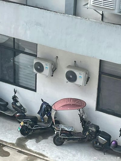 这是一张实拍的户外场景图，建筑外墙上挂着两台美的品牌的空调外机，下方地面整齐停放着四辆电动自行车，其中一辆还加装了粉色遮阳棚。巧合的是，两台空调外机的出风口正好对着下方的电动车，视觉上仿佛是在给电动车“吹空调”，形成了充满趣味的生活化巧合场景，让人忍俊不禁。