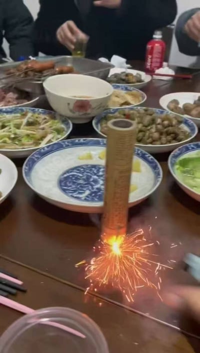 这张图呈现了一个十分荒诞的聚餐场景：深棕色的木质餐桌上摆满了各式各样的家常菜，包含炒菜、卤味、点心、汤碗等丰富餐食，桌子后方露出几名聚餐人员的上半身，有人正举着玻璃杯似乎在敬酒。而餐桌靠前的位置立着一枚已经被点燃的窜天猴类烟花，烟花底部正迸射出明亮的火花，随时可能向上飞窜，本该温馨热闹的聚餐氛围和极具危险性的放烟花行为形成强烈反差，充满离谱的搞笑感。