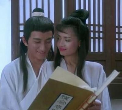 图中是1991年香港古装电影《玉蒲团之偷情宝鉴》的剧照，画面里吴启华饰演的未央生与叶子楣饰演的艳芳身着素净白色古装，二人紧密依偎在一起，共同低头翻看手中的线装古籍，脸上都带着柔和的笑意，氛围暧昧缱绻。背景是中式传统木质窗棂，为画面增添了古典雅致的氛围，精准呈现出影片中男女主角缠绵互动的场景。