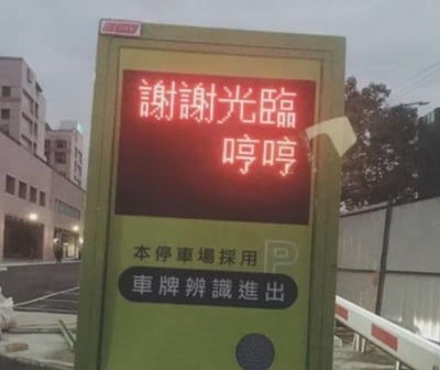 这是一张拍摄于停车场出口的设备界面照片，设备的电子显示屏上显示着“谢谢光临 哼哼”的文字，原本正式的送客语加上“哼哼”这个带有小情绪的可爱语气词，形成了强烈的反差感，让原本刻板的公共提示变得生动有趣。设备下方还标注了“本停车场采用 车牌辨识进出”的说明文字，背景能看到停车场外的建筑物和围栏，整体场景是真实的停车场出口环境。