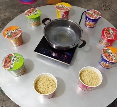 这张图呈现了一个极具共鸣的搞笑生活场景：圆形餐桌中央摆放着电磁炉与空锅，周围环绕着多桶不同包装的康师傅香辣牛肉面，还有两个已拆开、仅装着面饼的杯面，将泡面摆出了吃火锅的阵仗，精准调侃了过年期间家里来客但自己不会做饭的窘迫处境，用敷衍又幽默的方式化解待客难题，充满接地气的生活趣味，戳中不少不会做饭的人的痛点。