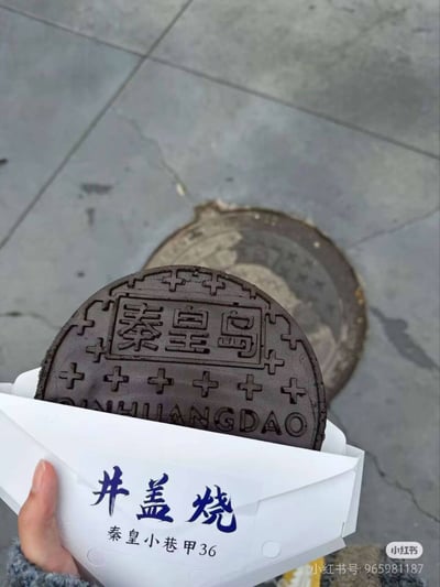 一只手握着印有“井盖烧 秦皇小巷甲36”字样的白色纸盒，盒内装着一块造型高度模仿井盖的圆形点心。点心表面清晰雕刻“秦皇岛”及“QINHUANGDAO”字样，边缘装饰十字花纹，与背景地面上的真实井盖形成趣味对照。整体以俯视角呈现，突出点心对井盖细节的逼真还原，展现了地方特色美食的创意设计巧思。