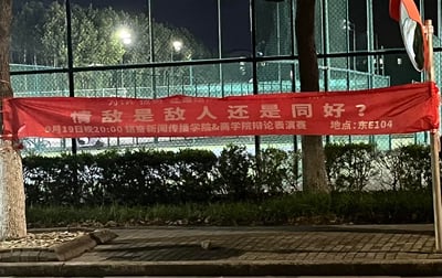 这是一张夜晚在大学校园拍摄的照片，画面中绿色的运动场地围栏前悬挂着一条红色宣传横幅，横幅上是新闻传播学院与商学院联合举办的辩论表演赛信息，核心议题为「情敌是敌人还是同好？」，比赛时间为9月19日晚20:00，地点在东E104。这个贴近年轻人情感生活的趣味议题，很容易引发学生的兴趣与讨论。