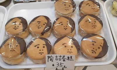 图片展示了一个白色托盘，内有8个独立包装的圆形糕点。每个糕点顶部用巧克力酱绘制了类似人脸的图案：左侧有深色“头发”，面部有简单勾勒的眉毛、眼睛、鼻子和略带愁容的嘴巴，整体神似歌手陶喆的面部特征。托盘前方放置一张手写价格牌，内容为“陶喆蛋糕 35元”。糕点疑似夹心蛋糕，包装透明可见内部奶油夹层，背景显示为 bakery 陈列柜局部，侧面有其他食品托盘边缘。图片通过将糕点外形与明星面部特征结合，形成幽默有趣的视觉效果，吸引顾客注意。