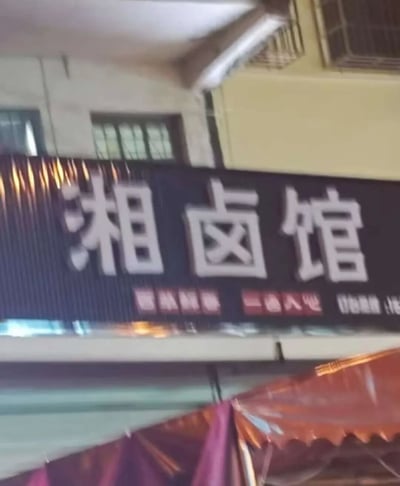 图片展示了一家名为“湘卤馆”的餐馆招牌实拍场景。招牌为深色底，上方有醒目的白色大字“湘卤馆”，下方配有模糊的红色和白色小字（疑似宣传语或营业时间）。背景可见建筑物的窗户、金属格栅及空调外机，底部有红色与紫色相间的遮阳篷，整体呈现出街头小店的日常氛围。