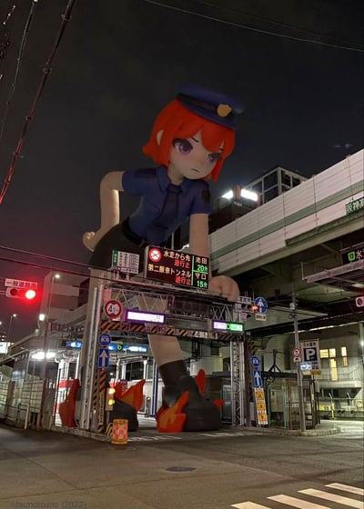 这是在日本大阪的夜间城市街头，一个巨型卡通女警立牌被设置在道路限高架旁，借助视觉错位效果，仿佛女警正叉腰扶着交通指示牌，脚边的火焰装饰增添了二次元感。背景是城市高架桥、交通信号灯与实时道路通行指示牌，指示牌上标注着水走、池田、守口方向的道路限行与等待时间信息，原本单调的交通场景被赋予了新奇的二次元趣味，让人眼前一亮。