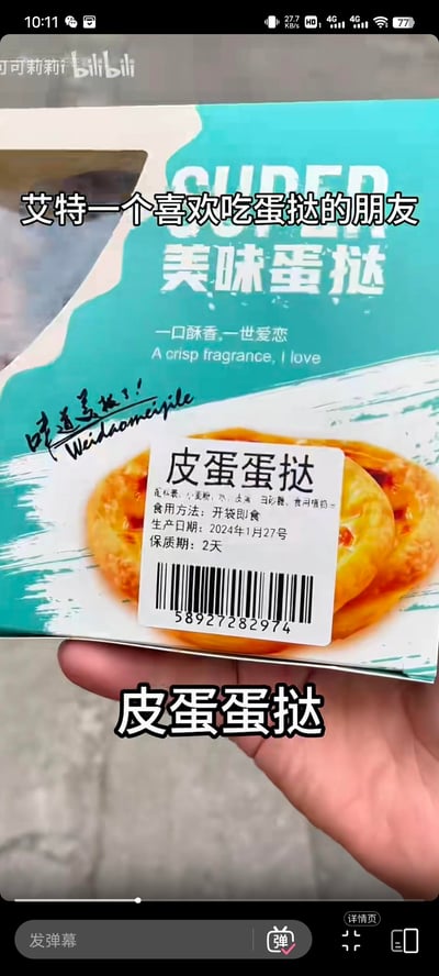 这是一张B站短视频的截图，展示了一个被恶搞改造的蛋挞包装：原本的「美味蛋挞」包装被贴上了「皮蛋蛋挞」的标签，配料表中加入了皮蛋，将常规蛋挞改造成了充满反差感的「黑暗料理」。图中配有文字「艾特一个喜欢吃蛋挞的朋友」，用来调侃好友，把这款奇怪的恶搞美食分享给对方，制造出搞笑的整活效果。