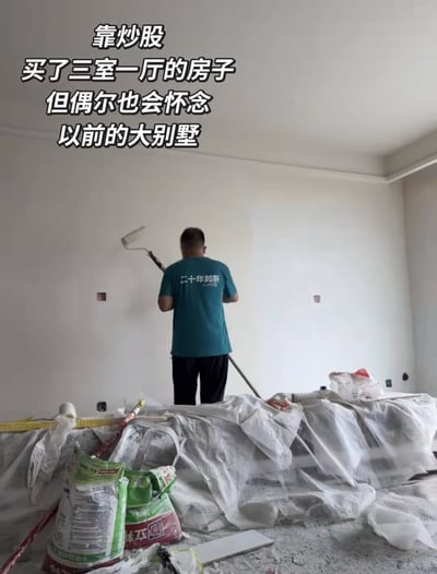 这是一张图文搭配的网络梗图，画面为正在装修的毛坯房室内场景，一名背对镜头的男性穿着蓝绿色短袖，手持滚筒刷正在给墙面刷漆，地面铺有装修遮盖布，还摆放着涂料袋等装修工具杂物。画面上方叠加了四行带黑边的白色文字，文字内容与画面中人物干装修活的场景形成强烈反差，用夸张的反差感玩梗，调侃炒股出现大幅亏损导致资产缩水、生活水平下降的情况，带有炒股人群对投资失利的自嘲幽默感。