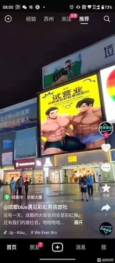 这是一张抖音APP的竖屏截图，展示了成都京都大厦外的城市广场夜景。广场上方的户外大屏投放着「成都遇见彩虹男孩旅社」的试营业广告，广告以明黄色为背景，主体是两名并肩微笑的卡通肌肉男性形象，右下角带有彩虹标识，明确指向LGBTQ+友好的定位，广告文字为「正在试营业 成都遇见彩虹男孩旅社 Meet Your Love in Chengdu」。屏幕下方的抖音界面显示，该视频由@成都blue遇见彩虹男孩旅社发布，定位为成都市京都大厦，配文写道「总有一天，成都的大街会到处是彩虹旗，还有我们的旅社名，哈哈哈哈...」，表达了对LGBTQ+文化在城市中更广泛传播的憧憬。视频的互动数据为：点赞7、评论2、转发1，BGM为《Up - KiiiU_ If We Ever Bro》，截图右下角带有「地饺吧」的水印，表明该截图可能来自贴吧分享。广场上有行人走动，整体氛围轻松，传递出社群文化的归属感与对未来的期待。