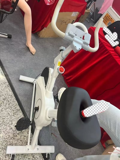 图片中展示了一台设计奇特的健身自行车，其最引人注目的特征是座椅部分采用了类似马桶座的设计，中间有一个明显的大洞。自行车主体为白色，位于一个铺有灰色地毯的展示区域。背景中可以看到一个人体模型的腿部，红色的桌布，以及一些纸箱，暗示这可能是在一个商店或展览场所拍摄的。图片右侧和底部有部分像素化处理（ checkerboard 图案），可能是为了隐藏某些信息或商标。这种将健身器材与马桶座设计结合的产品看起来非常滑稽和不实用，给人一种荒谬可笑的感觉。