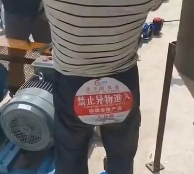 图片中展示了一位身穿灰白条纹上衣、深色裤子的打工人背影，其腰部后方醒目地贴着一张圆形红白相间的标识，上面印有“禁止异物进入”和“检验合格产品”字样。背景环境显示为工厂车间，可见灰色电动机等工业设备，整体场景充满生活化的职场幽默感，标识错位贴在人身后形成了意外的搞笑效果。