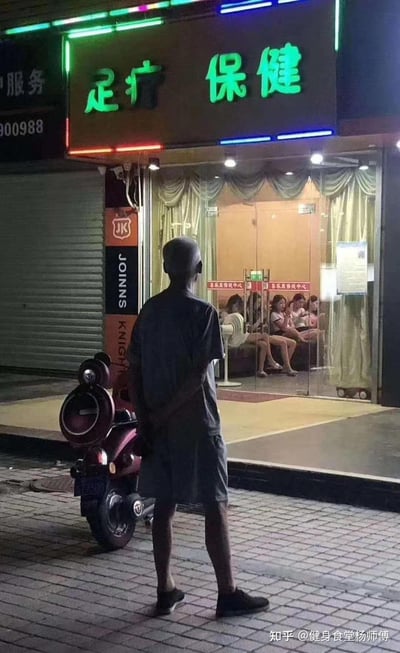 深夜的街头，一位光头大爷背着手站在一家亮着绿色发光招牌的「足疗 保健」店铺门前，透过玻璃门能看到店内坐着数位年轻女性，大爷背对镜头，似乎在观望店内的情况。这个充满市井气息的场景自带诙谐感，容易引发网友的调侃与遐想，场景本身无特殊双关或谐音梗，主要靠画面的反差感带来趣味。