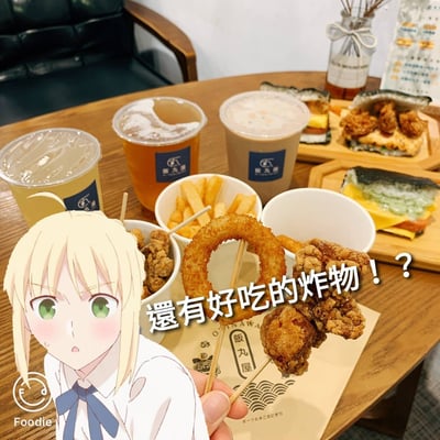 图片展示了一张木质餐桌上摆满美食的场景，包括三杯印有“飯丸屋”标志的饮品（疑似奶茶和茶饮）、薯条、炸鸡块、洋葱圈以及寿司类食物。一只手正拿着串有洋葱圈和炸鸡块的竹签。前景中叠加了一位金发绿眼的动漫少女形象，她面带惊讶表情，旁边配有文字“還有好吃的炸物！？”，整体营造出可爱又诱人的美食分享氛围，是真实食物摄影与动漫元素结合的创意图片。