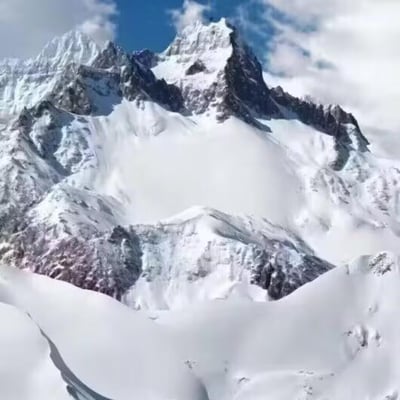 这是一幅展现高海拔雪山风貌的画面，巍峨的山峰被厚重的积雪与冰川覆盖，尖锐的峰顶刺破蓝天，部分深色的裸露岩壁与洁白的冰雪形成鲜明对比，云层在山巅缭绕，整体场景壮阔冷峻，充满了自然的雄浑气势。图中没有双关、谐音梗或暗示类内容。