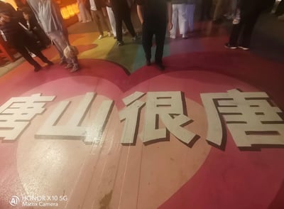 这是一张现场实拍的静态照片，拍摄于有灯光照明的场所，地面绘制有两个交叠的粉色爱心图案，爱心区域内印有带深色阴影的白色立体大字“唐山很唐”，文字后方有多名路人站立停留，画面左下角带有拍摄设备荣耀X10 5G的相机水印。